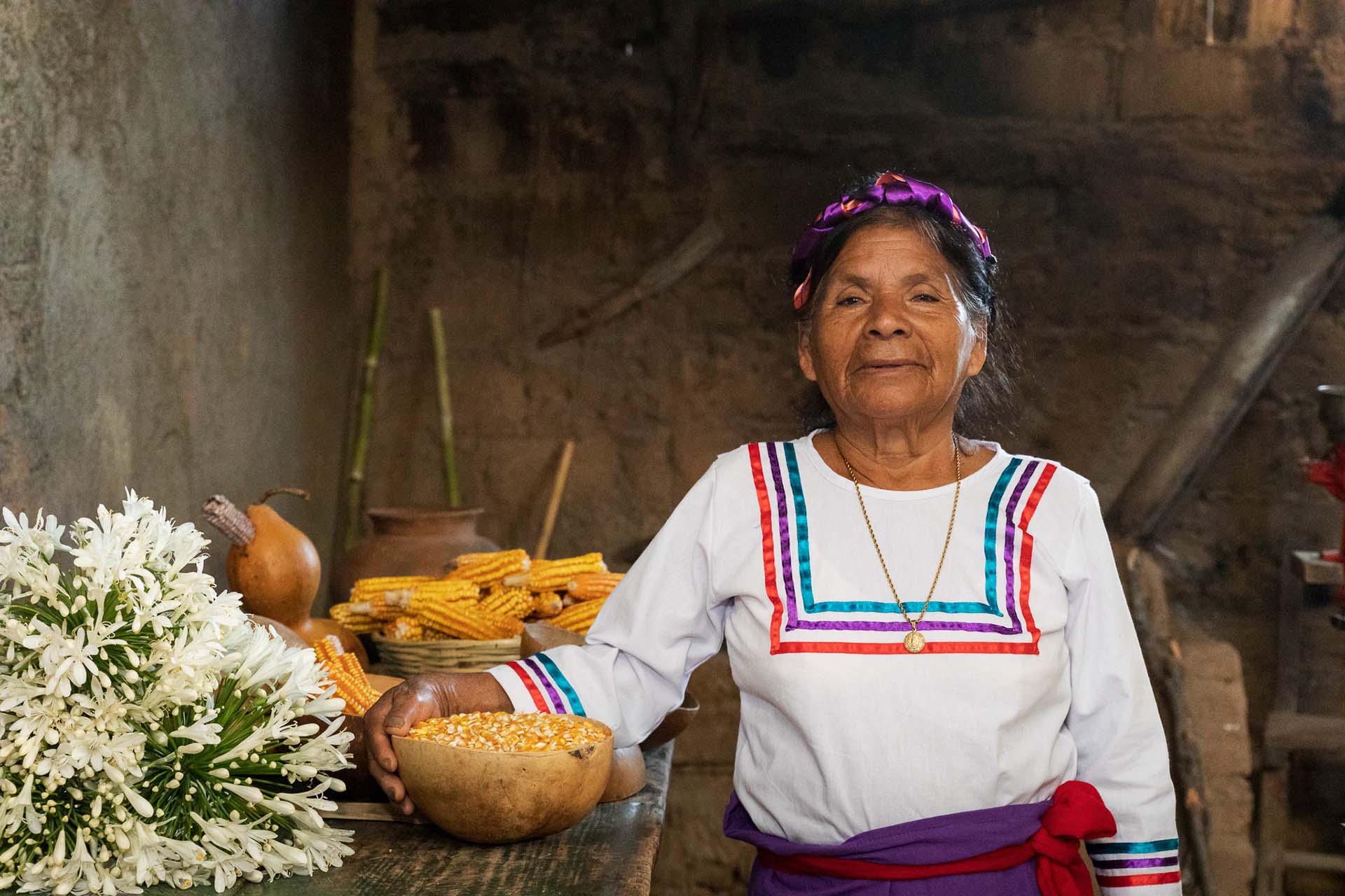 Doña Antonia, mujer chontal, posa sonriente en su cocina tradicional junto a mazorcas de maíz, flores blancas y una jícara con granos.