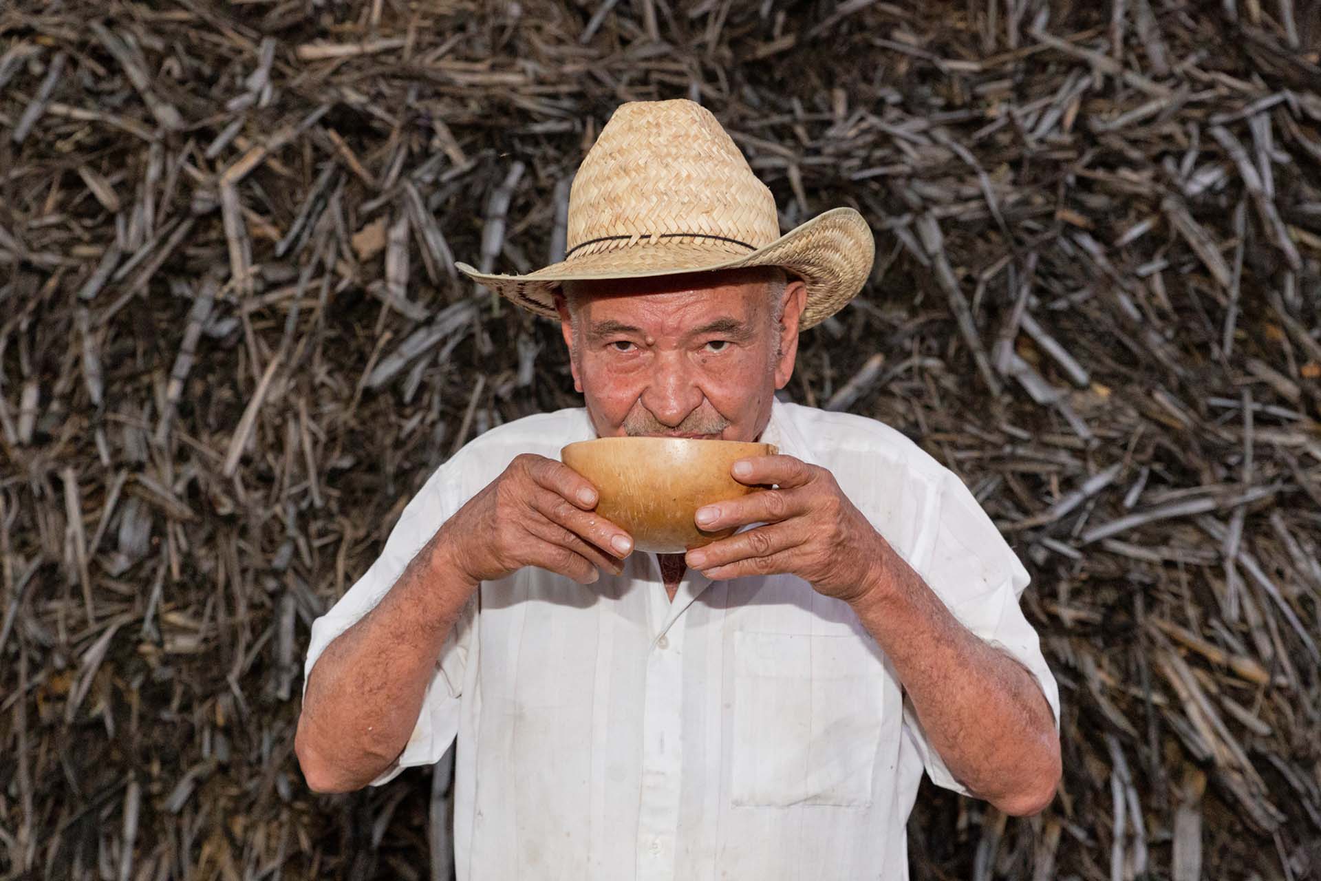 Don René, campesino oaxaqueño, bebe tepache de frutas en una jícara tradicional, frente a un fondo de bagazo de caña. Esta bebida es una de las 87 recetas documentadas en el libro Bebidas de Oaxaca, disponible en versión digital y física.