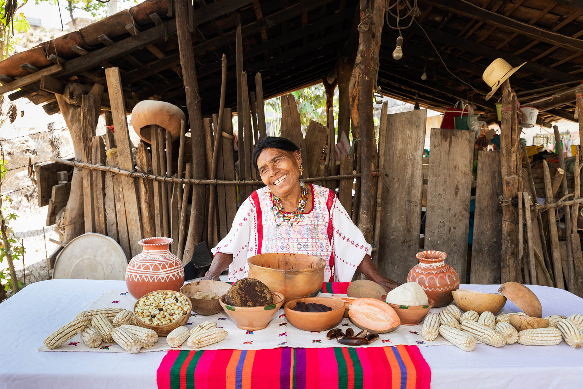 Mujer sonriente con vestimenta tradicional mixteca, sentada frente a una mesa con mazorcas, jícaras, cacao y utensilios de cocina, en una cocina de leña al aire libre en Oaxaca.