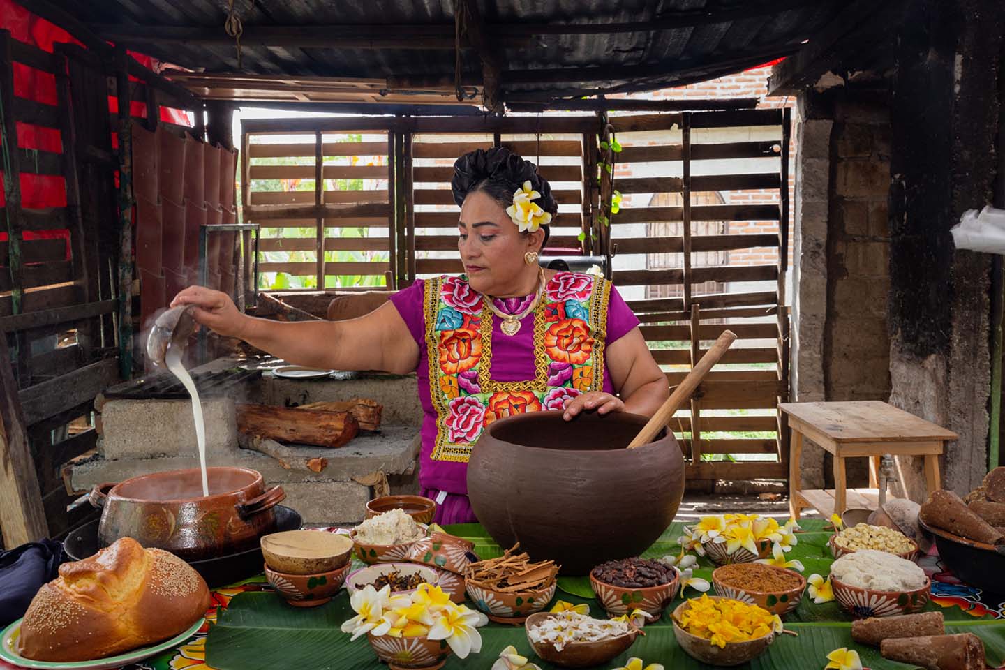 Una mujer istmeña, vestida con traje tradicional, vierte atole en una olla de barro rodeada de ingredientes sobre una mesa decorada con flores y hojas de plátano, durante la preparación del bupu.