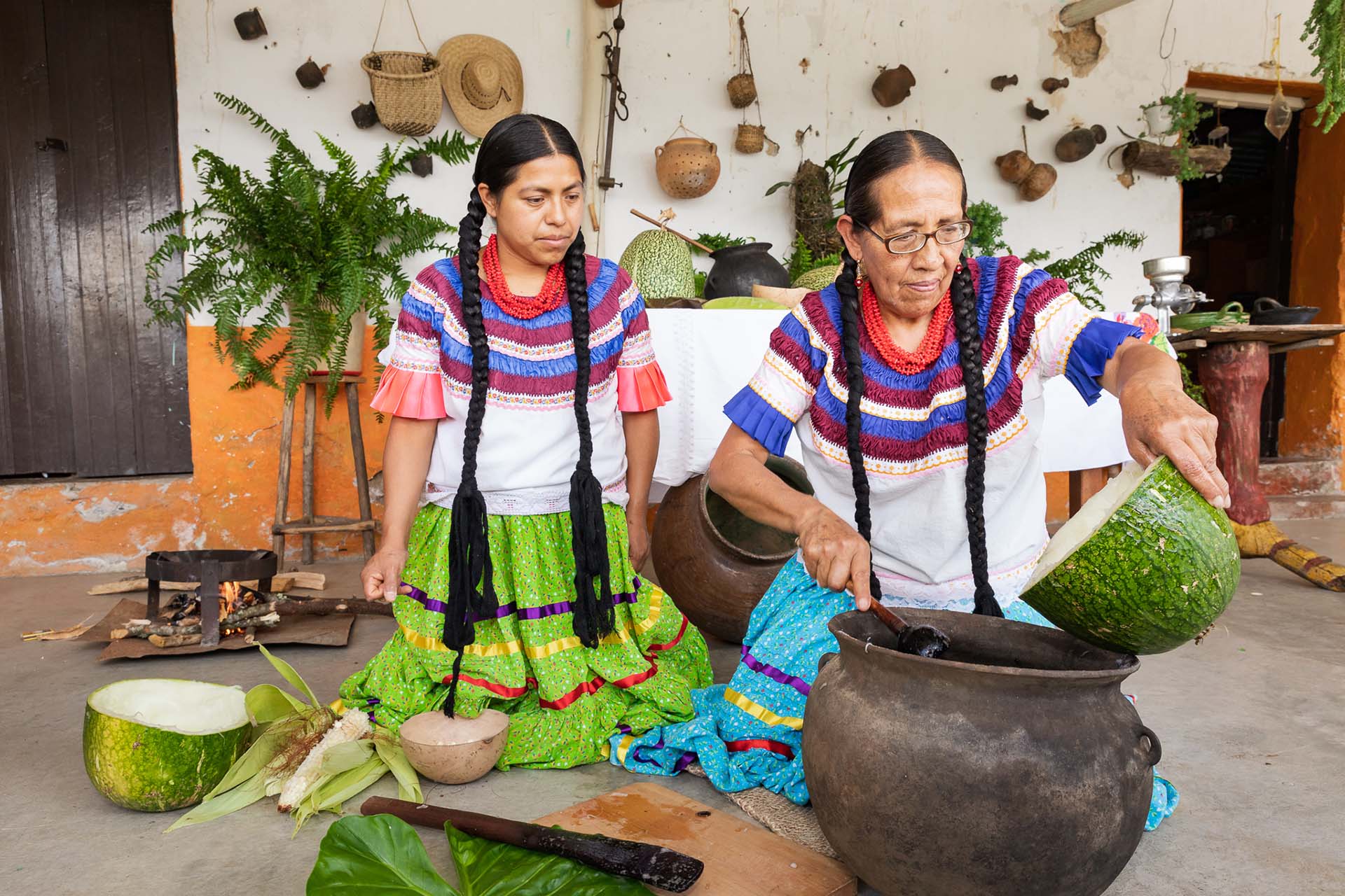 Doña Rosaura y su hija Marta, vestidas con ropa tradicional, preparan una bebida con chilacayota en una olla de barro sobre el piso de su casa en San Jerónimo Tecóatl, Oaxaca.