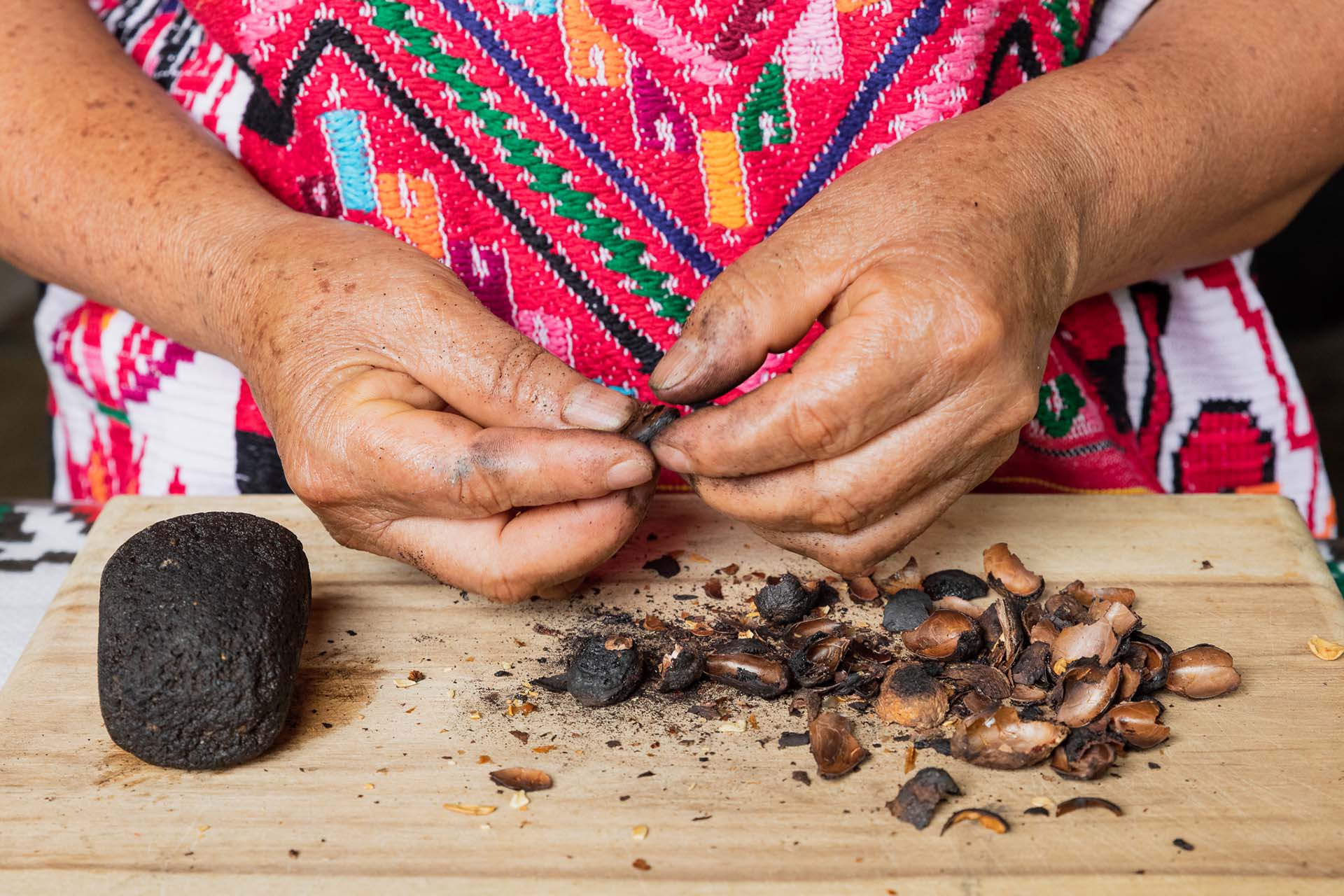 Manos de una mujer chinanteca pelando semillas de cacao tostado sobre una tabla de madera. A un lado, cáscaras vacías y una bola de masa negra de cacao. Lleva un huipil tradicional bordado en tonos rosas y rojos.