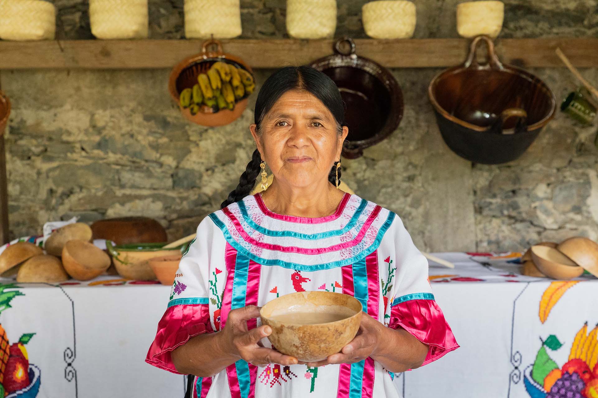 Retrato de Doña Emma Méndez García, originaria de Huautla de Jiménez, sosteniendo una jícara con atole agrio frente a una mesa tradicional con utensilios y bordados de la región