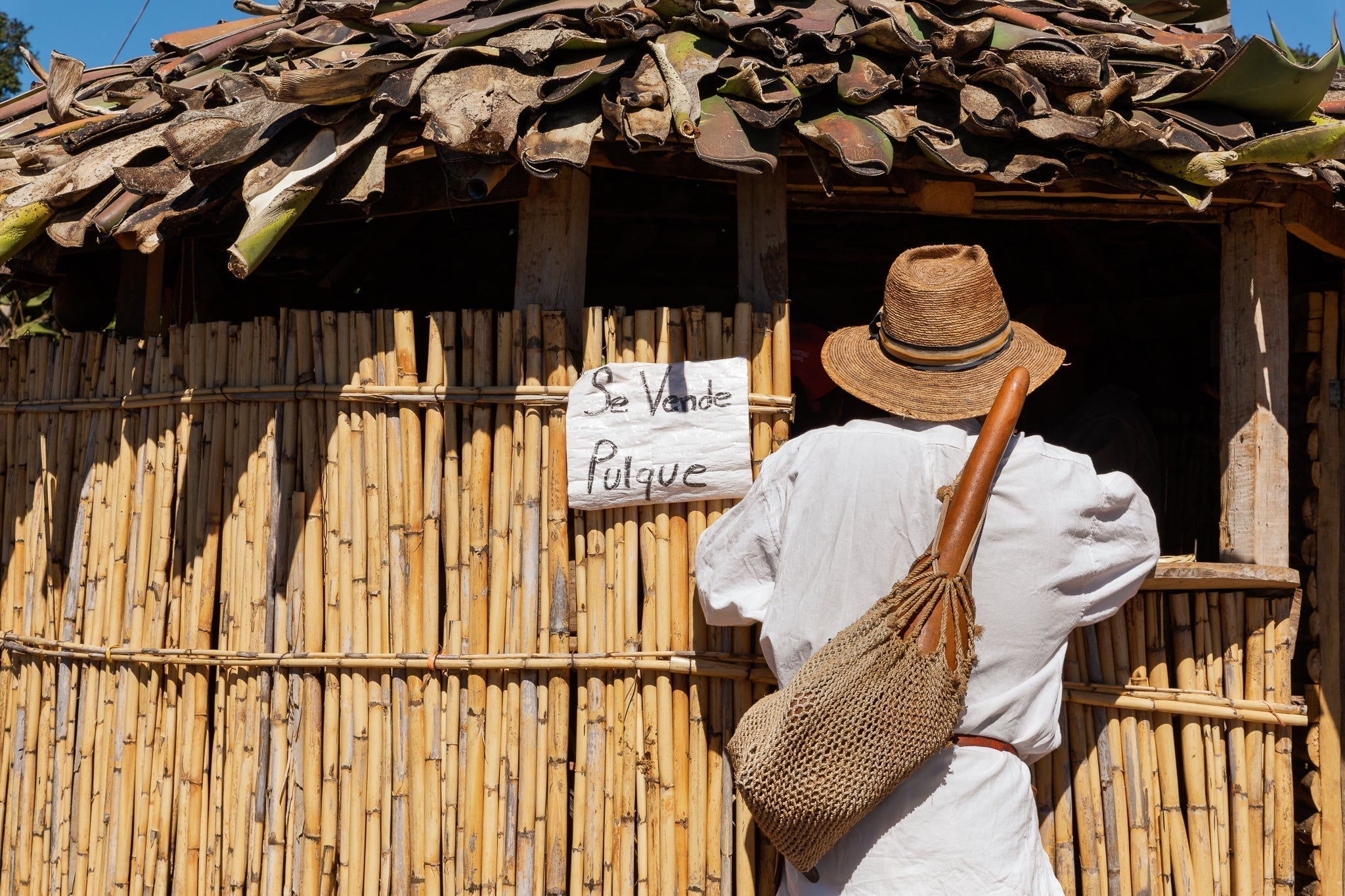 Persona de espaldas con sombrero y morral de ixtle, observando un letrero que dice “Se vende pulque” en una choza de carrizo con techo de hojas, en Nochixtlán, Oaxaca.