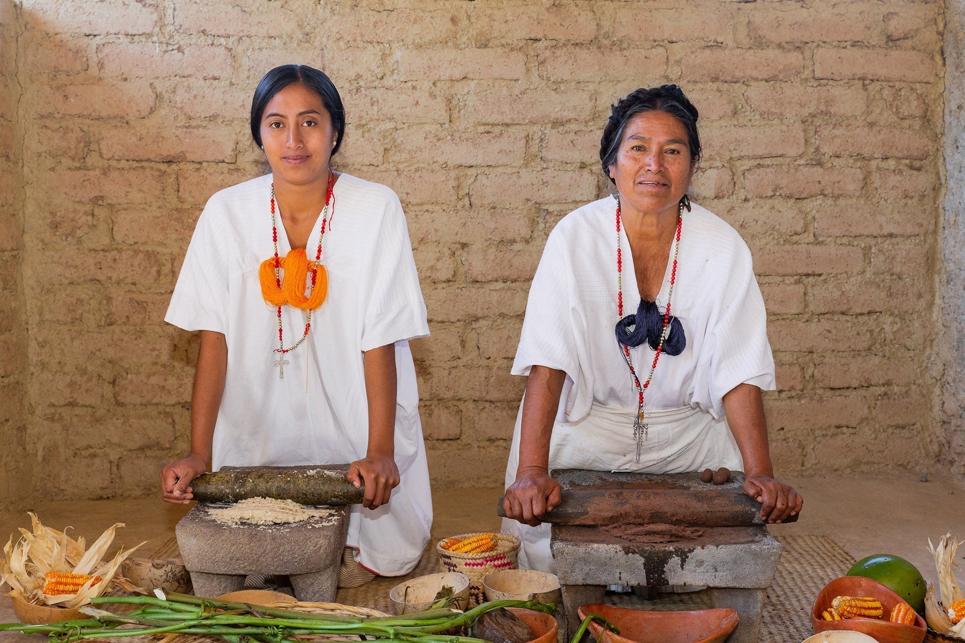 Dos mujeres con ropa tradicional blanca, sentadas frente a metates sobre petates, en una cocina de adobe, rodeadas de ingredientes para preparar pozontle.